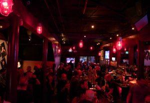 Interior of a crowded gay bar in Castro, San Francisco. Red lights illuminate the space above patrons at the bar. Gay travel.