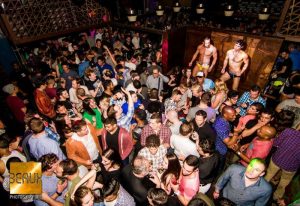 Crowded Beaux gay bar in San Francisco. Two shirtless dancers on stage. A diverse crowd of men and women partying. Nightlife scene.