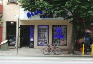 Blue Door Video, a gay sauna in New York. Storefront with blue neon sign. Bicycles parked outside. Gay travel destination.
