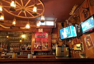 Interior of Branded Saloon, a gay bar in New York. Rustic decor with wagon wheel chandelier, lanterns, taxidermy, and TVs. A unique gay travel spot.