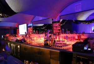 OMG Gay Bar in San Francisco: A long bar with bottles, glasses, and drink menus illuminated with red and blue lights. Nightlife.