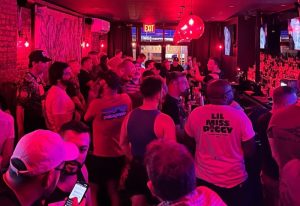 Crowded FLEX gay bar in New York City. Men socializing under red lighting. Nightlife scene, bar with patrons. Gay travel advice.