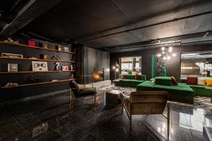 Empire Suites gay-friendly hotel lounge. Dark interior with a fireplace, green sofas, and shelves with decorative objects. Modern design.