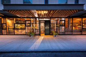 Kimpton Hotel Eventi, gay-friendly hotel entrance. Glass facade, lights, and plants. Welcoming ambiance for gay travelers. NYC hotel.