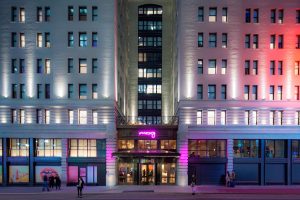 Exterior of Moxy NYC Times Square, a gay-friendly hotel. The building is lit with pink and red lights. Neon sign above the entrance.