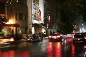 Night view of the Sofitel New York, a gay-friendly hotel. Flags of the USA and France fly above the entrance. Cars drive on wet street.
