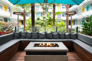 The Burrard Hotel's gay-friendly courtyard. Outdoor seating area with a fire pit, lush greenery, and colorful hotel balconies. A relaxing space.
