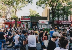 Good Judy Bar for queers in New York. Outdoor crowd scene, people gathered on the street in front of shops, including a laundromat.