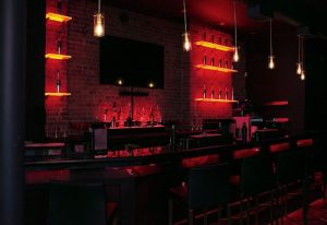 Lambda Lounge gay bar in New York. Bar with red lighting, brick wall, shelves with bottles, and hanging lights. Gay nightlife.