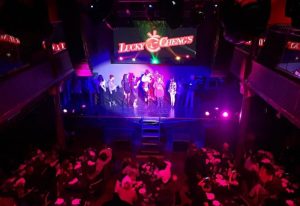 Lucky Cheng's Drag Cabaret in New York. Performers on stage under lights, viewed from above. Tables with patrons visible below.