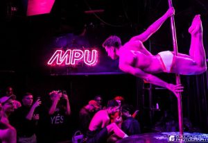 Le Belmont gay dance party in Montreal: A shirtless dancer performs on a pole under neon lights, entertaining a crowd.