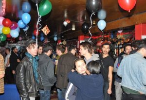 Inside Metropolitan Bar for Queers in New York, a lively crowd of gay men mingle beneath colorful balloons, enjoying drinks & camaraderie.