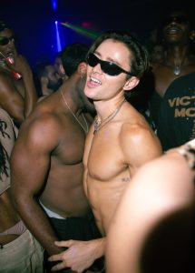 Gay New York: Men dancing shirtless at The Rosemont, a popular gay bar in NYC. A man in sunglasses smiles at the camera. Nightlife scene.