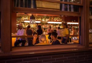 New York bar scene: View through window of people at a bar. Bottles line the shelves, lights hang above. Gay travel advice.