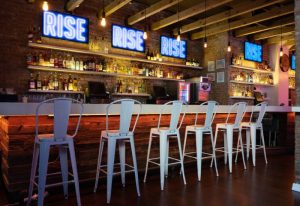 Rise Bar in New York: Interior view of the gay bar featuring white stools at the bar, neon signs, and a well-stocked liquor shelf.