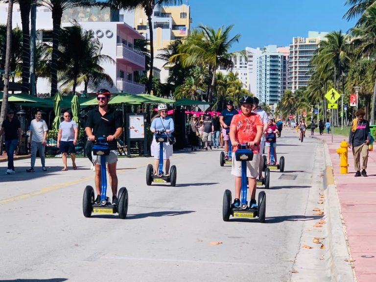 Gay travelers on Segways tour Miami's Art Deco District. Palm trees, colorful buildings, and sunny skies create a vibrant, welcoming scene.
