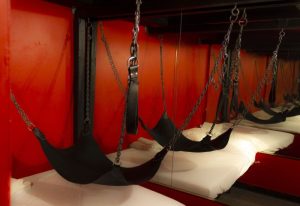 Toronto's Spa Excess: A bed with black sling, chains, and leather restraints. Red walls and mirrored background. Gay travel advice.