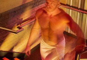 Man in towel at Steamworks bathhouse, a Chicago gay cruise club. He's on stairs, holding the rail. Red and yellow lighting.