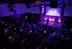 The Academy Social Club in San Francisco: Drag queen performing on stage for a gay audience. Purple lighting, disco ball, & cabaret vibe.