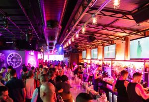 The Spot gay bar in New York. Crowded interior with men at the bar, TVs, and purple lighting. Gay travel advice.