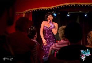 Drag queen performs at The Wiggle Room, a gay bar in Montreal. Live show with performer in purple sequin dress and gloves. Audience members watch.