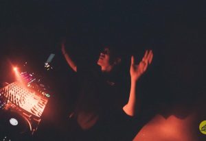 Gay travel: DJ with arms raised at a dark party. Nightlife scene with music and dance. Potent event for gay men.