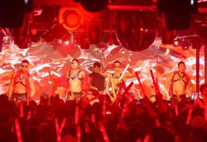 Gay travel: Men in harnesses perform at a club with red lighting and a disco ball. Crowd with red glow sticks. Destination nightlife.