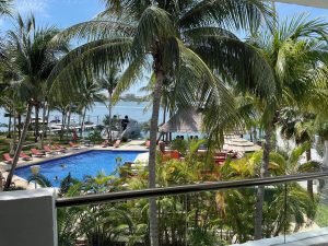 Sina Suites gay-friendly hotel view. Palm trees frame a pool with lounge chairs, a palapa, and ocean views. A relaxing tropical gay vacation spot.