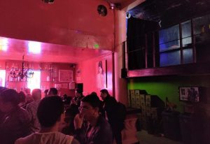 Interior of Marrakech Salon Gay Bar in Mexico. People socializing, bar with bottles, red lighting, and a mezzanine level. Gay travel advice.