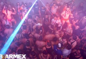 Overhead view of a packed Nicho Bears Bar gay party in Mexico. Men in harnesses dance under a light beam in a smoky, vibrant atmosphere.