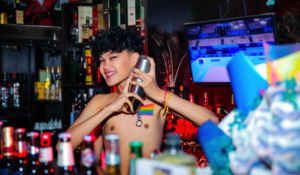 Shirtless bartender at POC Gay Bar in Phnom Penh, Cambodia. Rainbow flag necklace, shaker, and bottles. Gay travel.