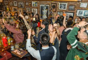 Polite & Co Hanoi: Gay men celebrate with champagne and confetti in a bar with framed photos. Gay travel advice for Hanoi nightlife.