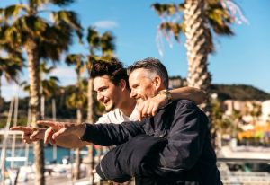 Father and son pointing at harbour. Gay travel in Cancun. Gay-friendly hotels. Palm trees, blue sky. Vacation, travel together.