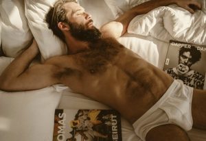 Bearded man sleeps in bed at a gay-friendly hotel. Male model in briefs, magazines nearby. Playa Del Carmen travel for gay men.
