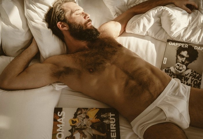 Bearded man sleeps in bed at a gay-friendly hotel. Male model in briefs, magazines nearby. Playa Del Carmen travel for gay men.