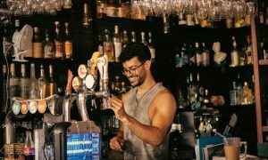 Bartender pouring beer at a Benidorm gay bar. Shelves of liquor bottles line the back. Nightlife scene, gay travel advice.