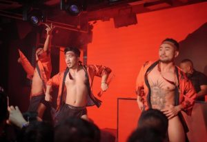 Destination: Asian male dancers perform at a Beijing gay club. Performers wear headbands, orange robes, and minimal clothing. Red lighting.