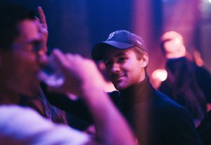 Man at a Gay Event in Paris. Nightlife scene with purple and blue lighting. The man wears a baseball cap and turtleneck. Paris gay travel.