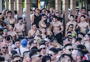 Crowd of shirtless Asian men with sunglasses at a gay travel event. Group of muscular men, some with tattoos, enjoying a pool party.