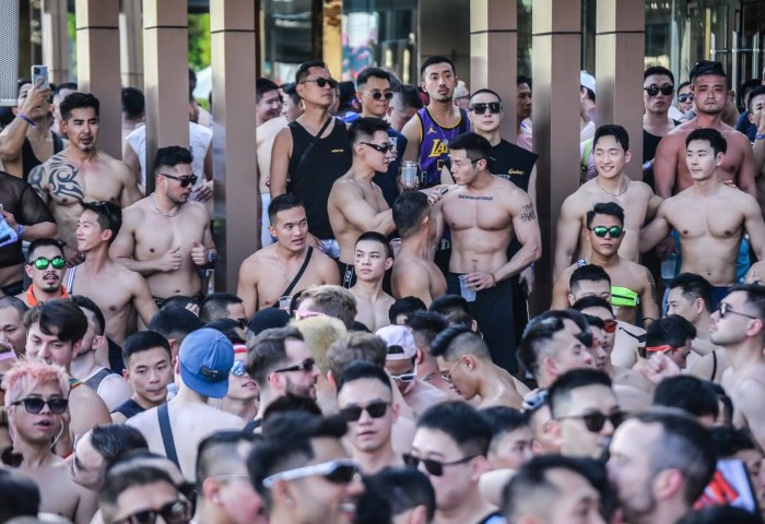 Crowd of shirtless Asian men with sunglasses at a gay travel event. Group of muscular men, some with tattoos, enjoying a pool party.