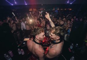 Two shirtless DJs at a Melbourne gay bar. Crowd dancing, lights, and music create a vibrant atmosphere. Gay nightlife scene.