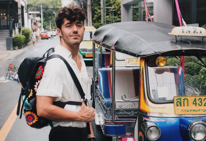 Gay traveler, Benjamin Thorpe, in Bangkok, Thailand. Holiday Inn Express Bangkok Sathorn in background. Tuk-tuk taxi and backpack.