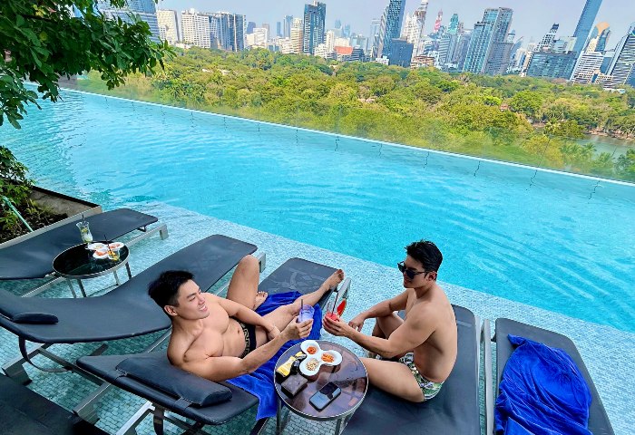 Gay couple at SO Bangkok hotel pool, Thailand. Men toasting drinks with city skyline view. Gay travel in Bangkok. NicHwan enjoying vacation.