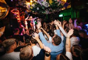 VIA, a gay event in Manchester. A performer on stage with a crowd of people raising their hands in excitement. Disco balls hang from the ceiling.