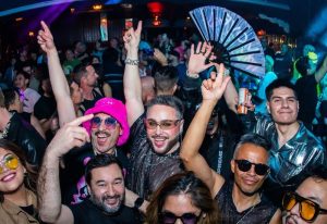 Gay club scene in Ibiza: Diverse group partying, hands raised, smiles, stylish outfits, fan, and colorful lighting. Ibiza gay nightlife.