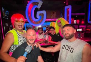Barcelona gay bar 700X480: Group of gay men and drag queens smiling at camera in a vibrant bar with neon lights and cocktails. Nightlife fun.
