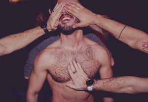 Man surrounded by hands at a Barcelona gay club. Shirtless, smiling man has his eyes covered and chest touched by other men's hands.