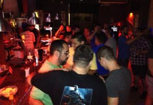 Inside Bacon Bear Bar, a gay bar in Barcelona. Men gather, some at the bar, others in conversation, creating a lively, social atmosphere.