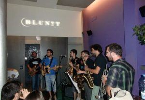 Live band at Blunit gay bar in Barcelona. Musicians playing instruments, including saxophone, guitar, and trumpet, for a lively crowd.
