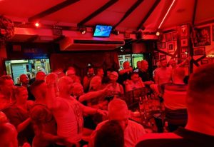 Centre Stage Bar, Gran Canaria. Red-lit interior of a gay bar, crowded with men. TV screen above the bar. Nightlife scene.